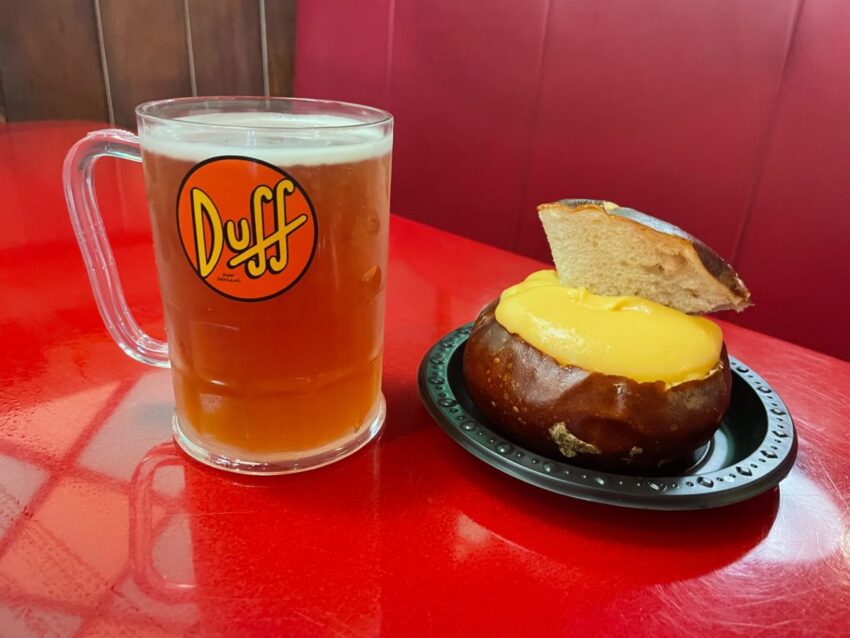 A glass mug of beer with a "Duff" label next to a bread bowl filled with cheese sauce and a bread piece on top, placed on a red table, perfect for celebrating Dufftoberfest.